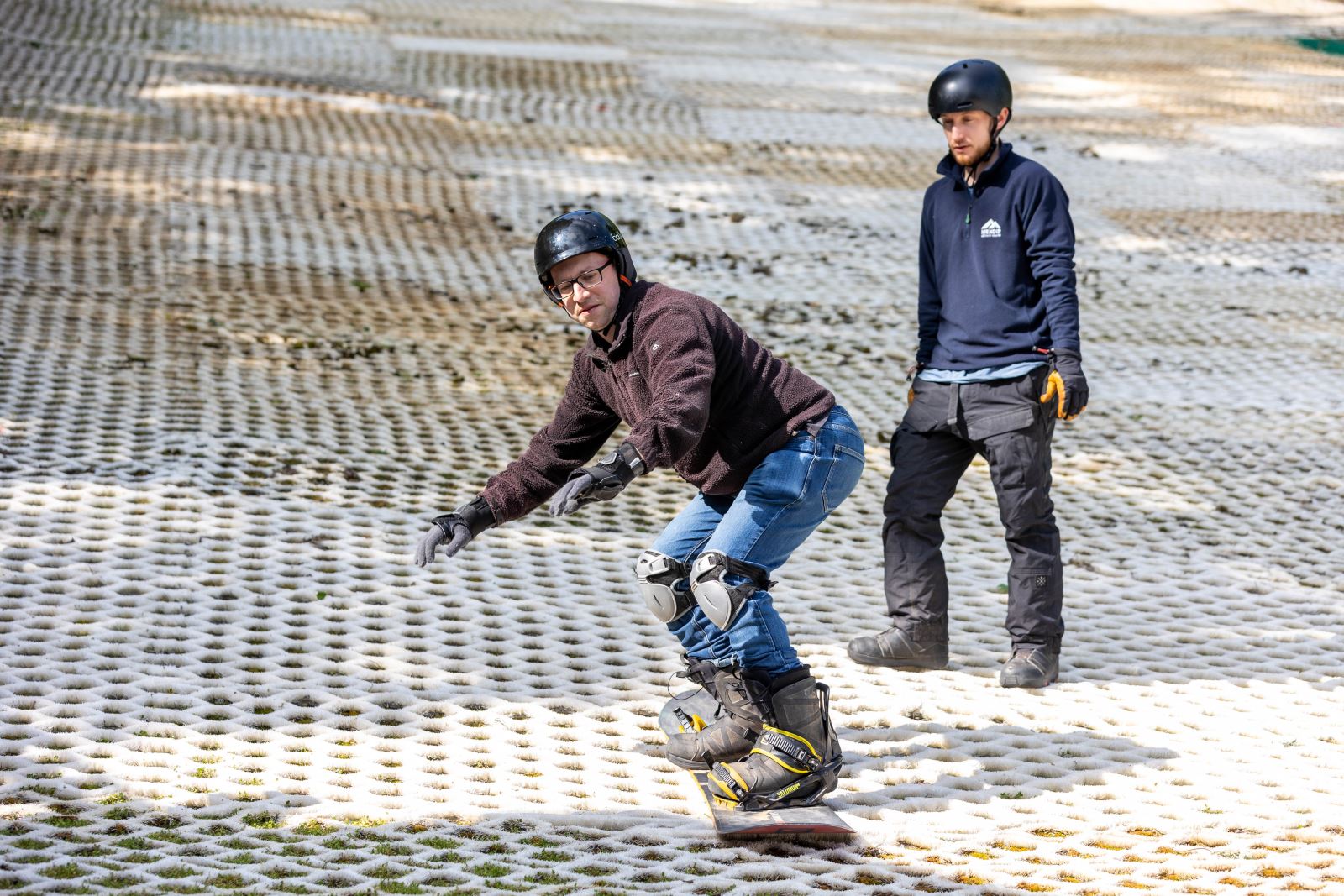 A man having a snowboarding lesson under the watchful eye of his male instructor on the dry ski slope at The Mendip Activity Centre