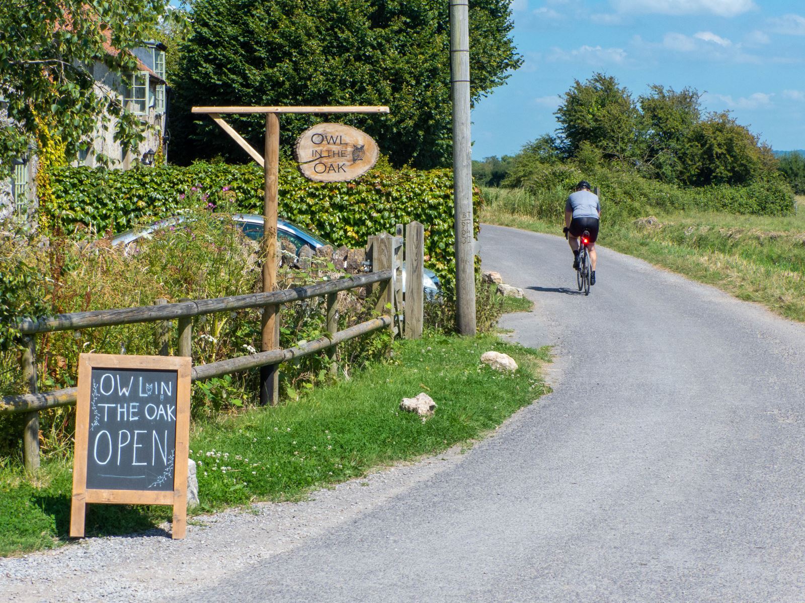 A cafe sign with a cyclist disappearing round the bend in the background