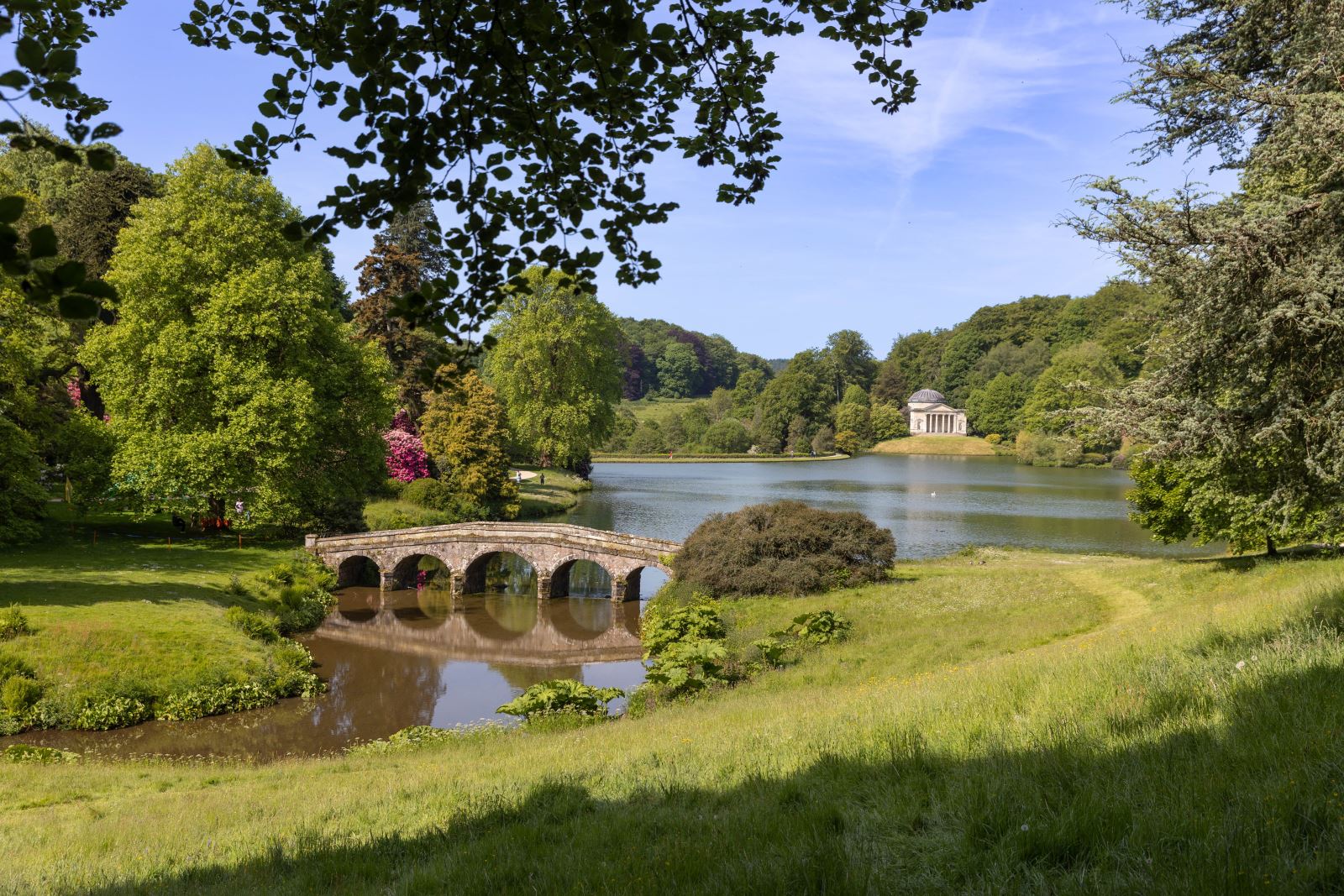 A fairytale view across a stone bridge and over the large lake at Stourhead to a pretty building with a round dome in the distance